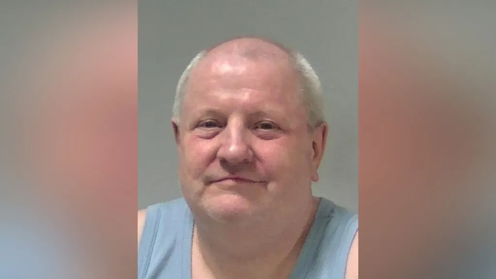 A custody photo of Steve Wright who is smiling at the camera. He is wearing a light blue vest and is bald with some grey hair around the side of his head.