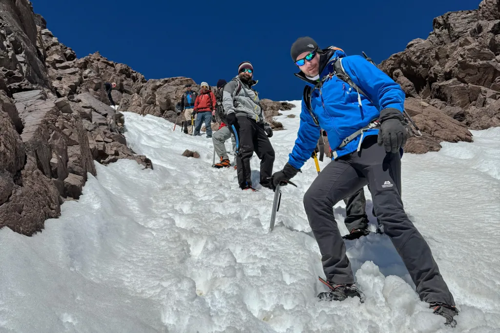 The photo is taken on the side of a snowy mountain, with some jagged rocks on show, and a deep blue cloudless sky. The photo has been taken from below with Gerwyn stood in the foreground of the photo, and five other men visible to varying degrees in the background. Gerwyn is leaning on an ice axe which has been wedged in the snow. Every man is wearing warm winter gear and sunglasses. 