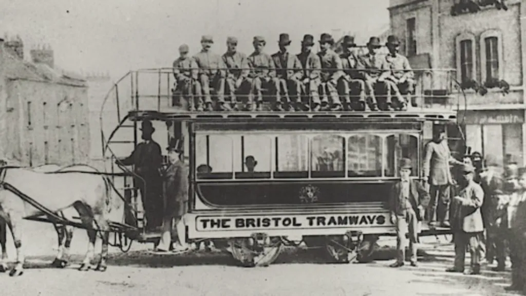Black and white image of the early trams in use in Bristol. Two horses stand at far left of the picture, at the front of the open-top carriage, with up to twelve people on board. 