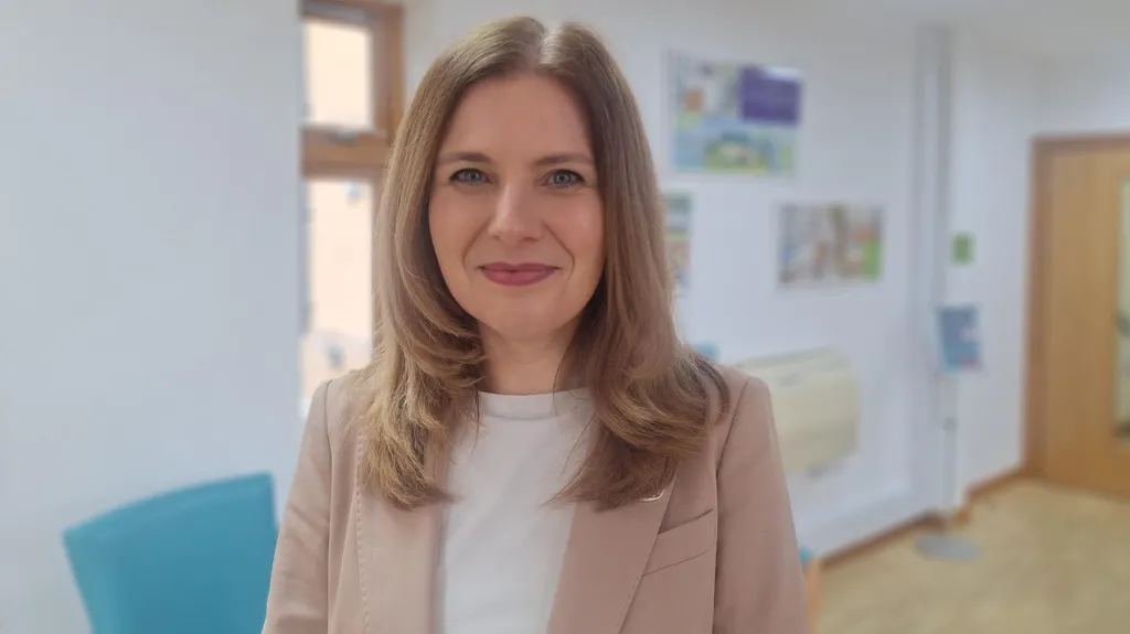 A woman stood in a corridor in a hospice. The background is blurred and she is in focus. She has shoulder-length blonde/brown hair and smiles at the camera. She has pink lipstick on and wears a beige jacket over a white t-shirt. It is a head and shoulders shot. 