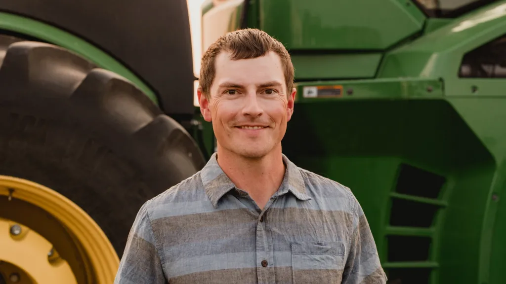 Smiling and wearing a stripey blue and grey shirt, Jake Leguee stands in front of large tractor.