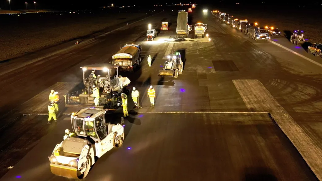 A still taken from a drone looking down at the runway at London Luton Airport. It is night and the runway is well lit with a series of construction vehicles in the foreground, including a roller and tipper trucks - one of which is tipping out material. The scene includes eight workers dressed in yellow high-visibility outfits. To the right of the picture is a long line of vehicles. 