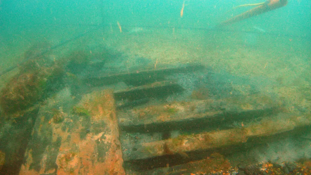 An underwater photo of a shipwreck. Timbers can be seen with algae growing on them. The water is green/blue and the wreck site on the seabed is marked out. 