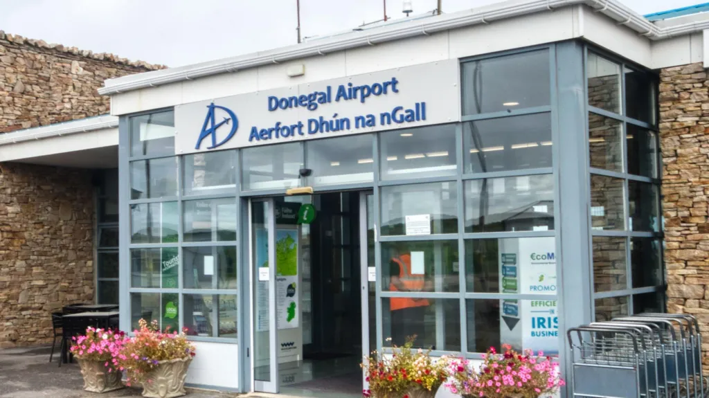 The front of Donegal airport, it is a small building with a glass entrance porch and stone walls on either side. The sign on the porch reads "Donegal Airport" in blue font on a grey background, with the Irish translation underneath. 