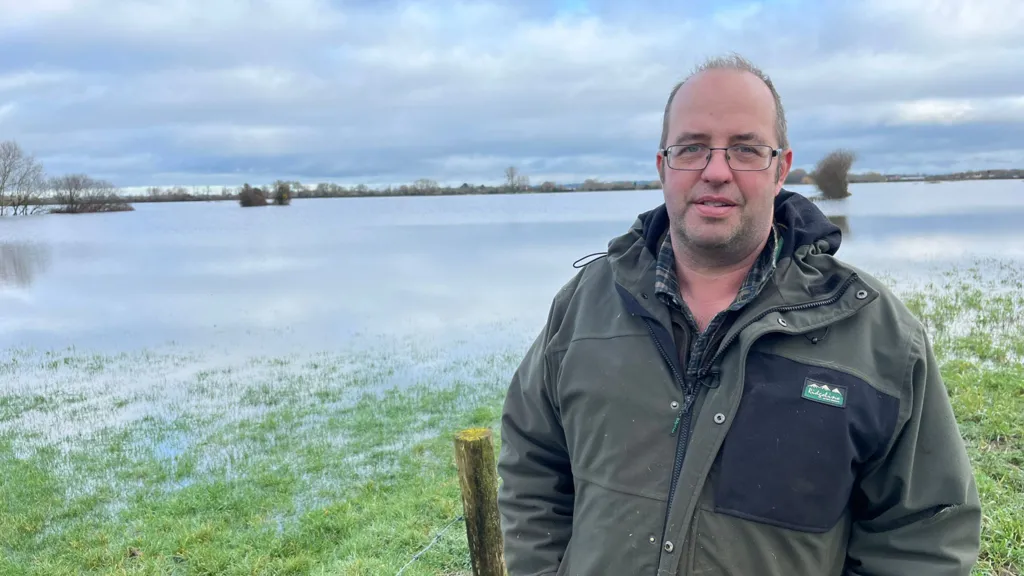 A man wearing a green jacket and checked shirt stands to the right of a grassy field which is submerged below flood water, the edge of a fence can be seen behind him. 