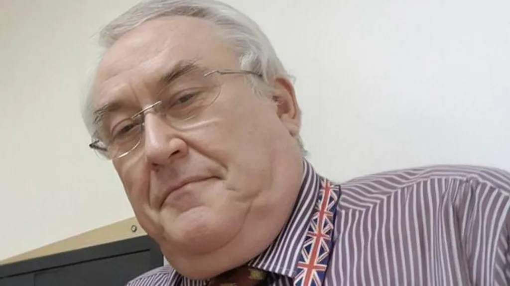 Leslie Irwin looking at the camera. He is an older man with grey hair and rimless glasses. He is wearing a purple and white striped shirt, a tie and a lanyard with a pattern of the Union Jack on.      