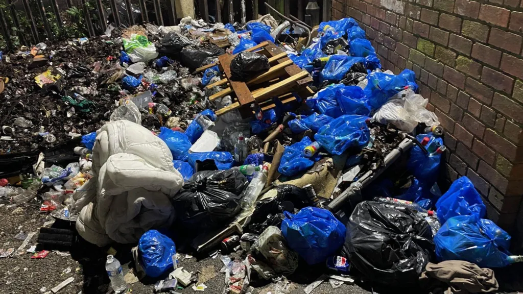 Bags of rubbish have been left beside bottles and drug needles at the corner of this car park