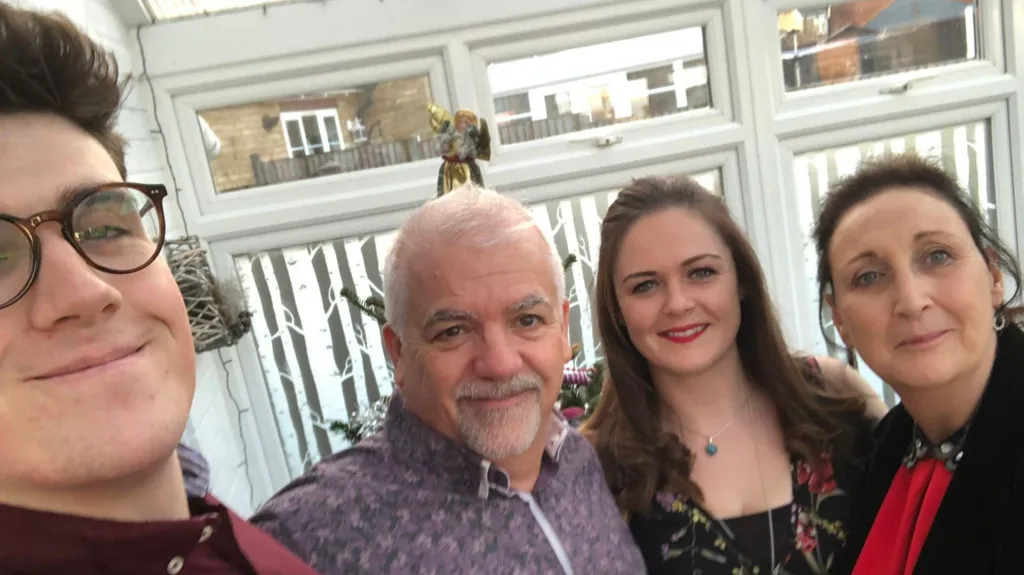 A selfie with four people in a row smiling at the camera. On the left is a man with brown hair and glasses wearing a burgundy shirt, then an older man with a grey beard and flowery purple shirt. On the right is a woman with long brown curly hair and red lipstick, and on the end is a woman with dark hair wearing a red dress.
