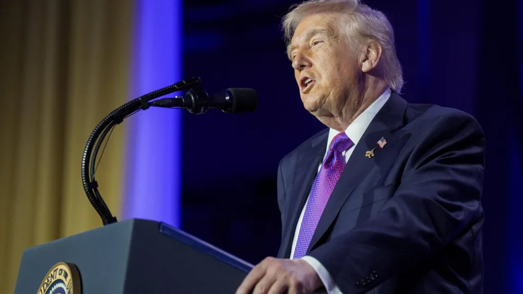 Donald Trump, who has light blonde hair and is wearing a dark navy blue suit over a purple tie and white shoot, speaks into a microphone on a lectern.