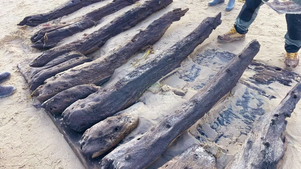The blackened timbers of a ship's hull can be seen on the shore, they are covered in sand. You can see three people's feet standing next to the section of wreck.
