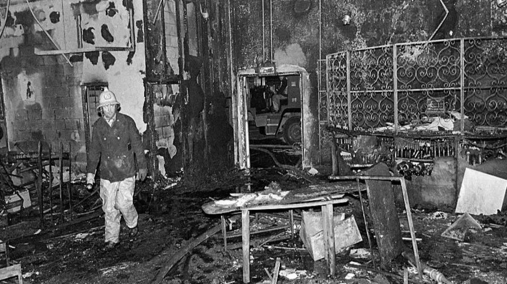 A black and white photo of a firefighter in the burned out wreckage of the Stardust nightclub in Dublin in 1981.  He is wearing a bright-coloured helmet and a protective uniform. The roof, walls and floor of the building have all sustained extensive fire and smoke damage. The remains of damaged tables and chairs are strewn around the floor. 