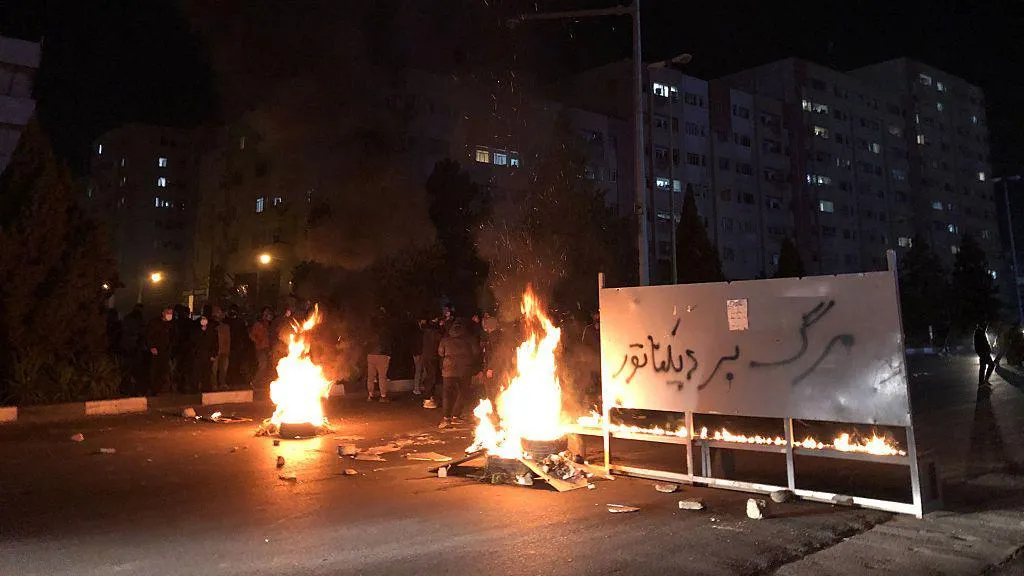 group of protesters in distance on street next to small fires and a sign that says "death to dictator"