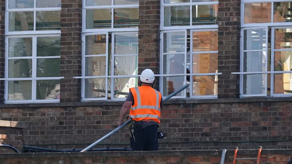 A construction worker in a hard hat and orange safety vest stands outside the brick building of a school, holding a long pole while working on or inspecting the exterior window frames. Several large windows reflect the surroundings, and scaffolding or support bars are positioned across them.