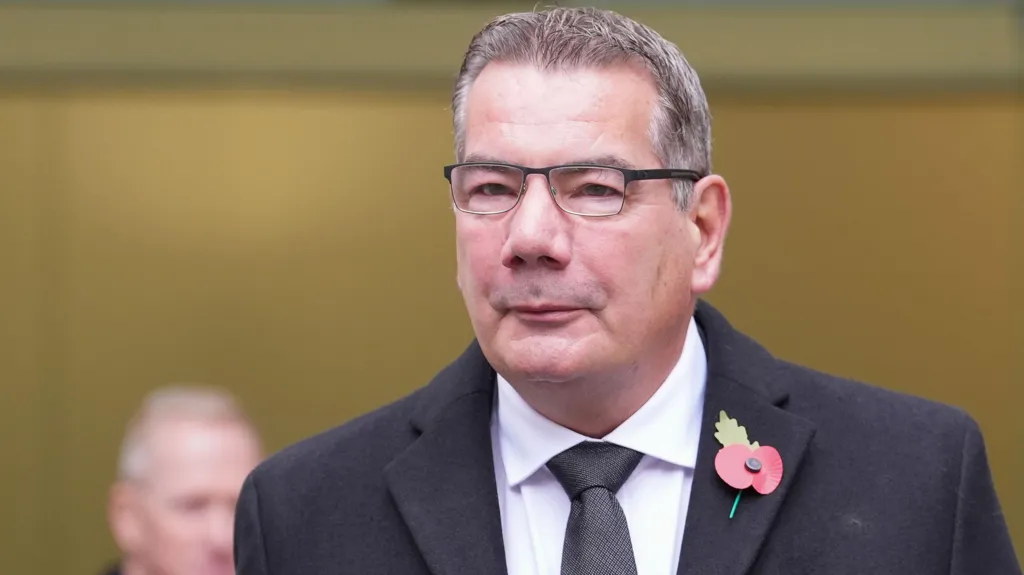 Nick Adderley walking near Westminster Magistrates' Court. He has short greying hair and is wearing glasses, a dark coat over a white shirt and black tie. On his left lapel is a poppy. 