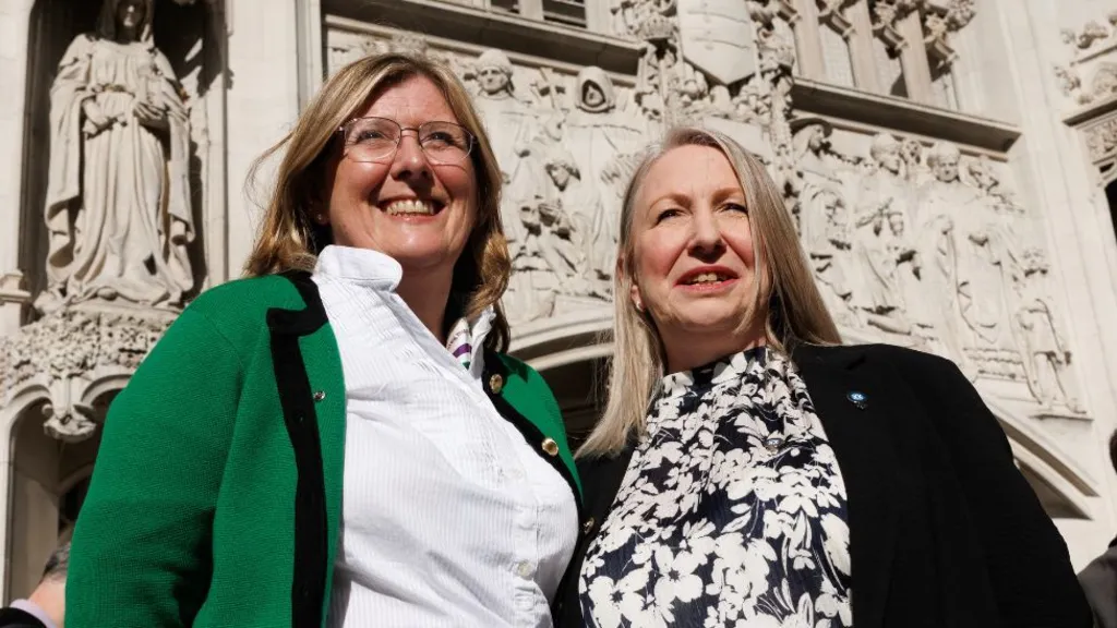Two women with fair hair outside a stone building with detailed carvings. They are photographed from a low angle, with the sun shining on their faces