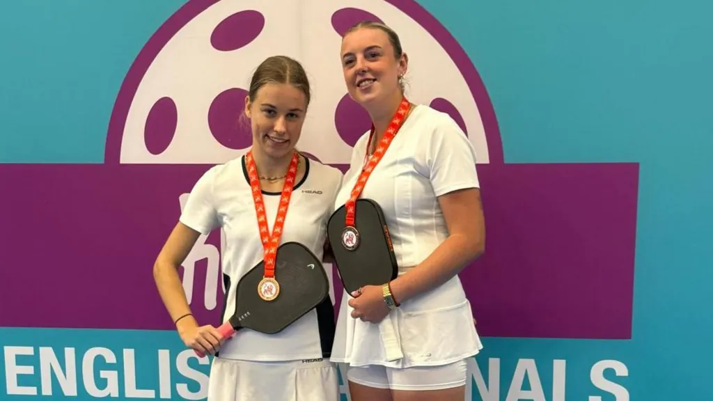 From left to right, Charlotte Pressley and Mollie Knaggs in autumn 2025 with their bronze medals. They both have light brown hair, pulled back from their face, are wearing white tops and shorts and holding up their paddles with their medals around their necks and the metal resting on the paddles. Behind them is a wall saying English finals in capitals. 