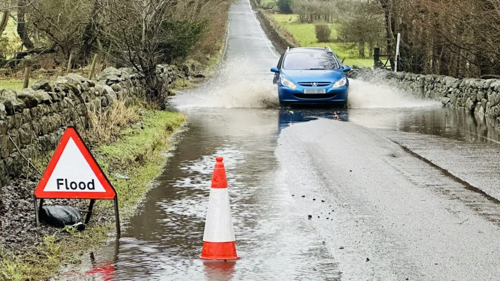 A blue car driving through a small flood in a country road. There is a hazard sign warning of the flood and a cone in some standing water.