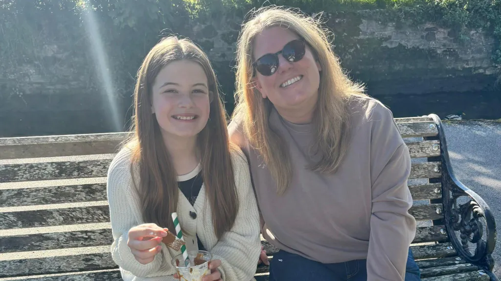 Rubie is sitting on a wooden bench with long brown hair and a cream cardigan. She is holding a drink with a green and white straw and is sitting beside her mum Sam smiling. Sam is wearing a light purple jumper and has long blonde hair and sunglasses. 