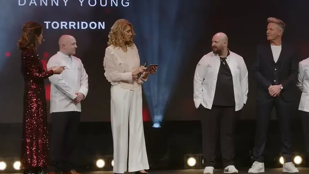 Presenters and chefs standing on stage during an award announcement at the Michelin Guide ceremonywith the names of recognised restaurants displayed on the large screen behind them. Danny Young, second from left, a bald man with white chefs jacket, stand between two presenters. Gordon Ramsey, wearing a dark suit with white t-shirt, stands at the right.