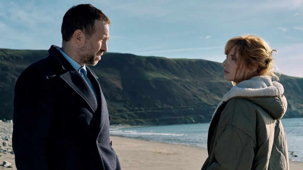 Reilly and Spall stand face to face with a beach in the background. Reilly is wearing a green winter coat, with her brown hair tied up. Spall is wearing a navy formal coat with a blue shirt, and has short dark hair and stubble.