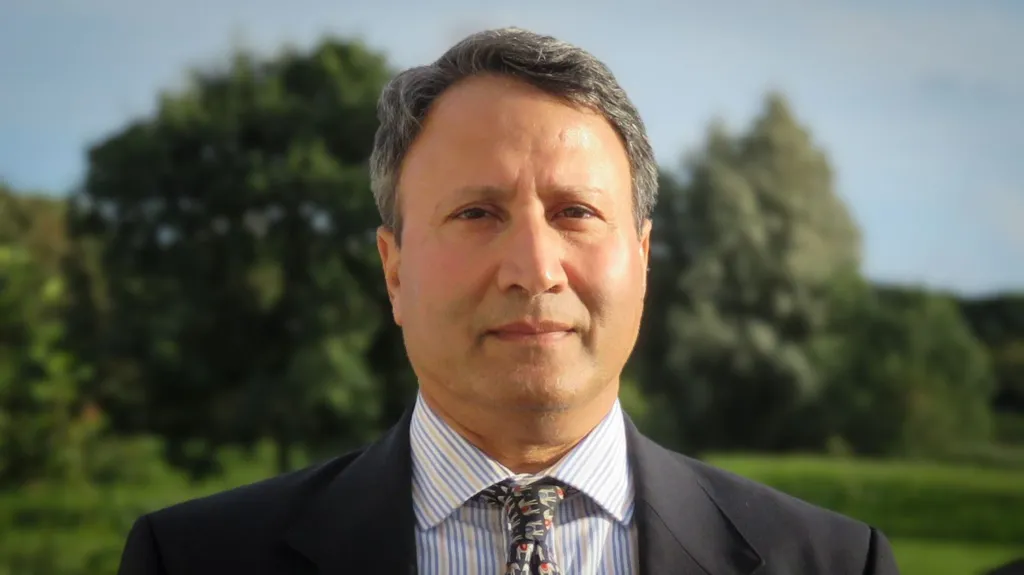 Rakesh standing outside, trees in the background, the sun is shining. He is wearing a dark grey suit, yellow and blue striped shirt and a tie with elephant pattern
