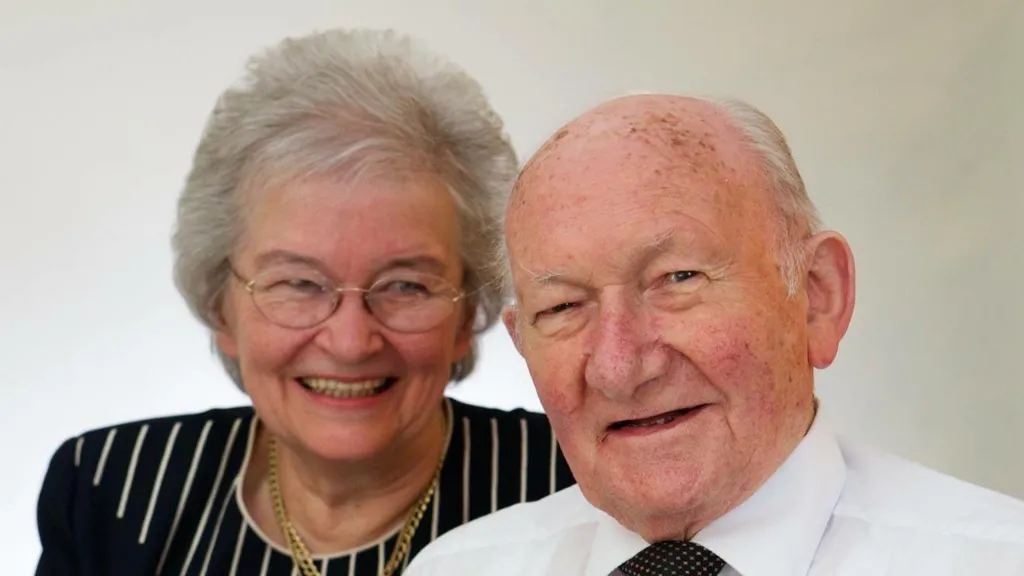 Janet McMillan with short grey hair, wearing glasses, a gold necklace and a black and white striped top, smiling to camera. She is to the right of a smiling Hammy McMillan. wearing a white shirt and black tie with short grey hair 