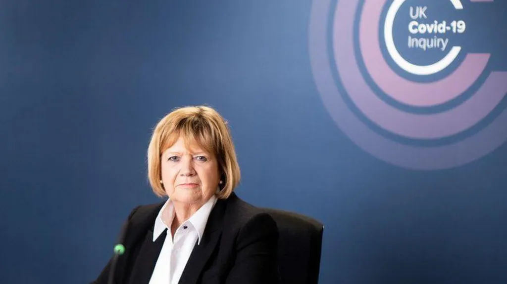 Baroness Hallett in front of a blue backdrop featuring the pink, purple and white Covid-19 inquiry logo. 