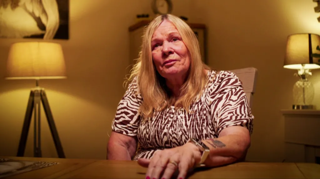 Cathy Broomfield is a woman in late middle age. She has dyed blonde chest-length hair. She is wearing an above the elbow sleeved dress with a brown and cream patterned dress. She is seated behind a table with her left hand resting on it. She wears an large square watch and a wedding ring. She has a tattoo on her lower left arm. She is sitting in a living room with a mantlepiece to her right with a lamp on it, and a standard lamp to her right. There is an out of focus cabinet behind her and a painting on the wall.