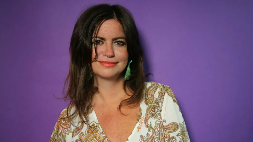 A picture of Dame Deborah James. She is smiling and wears a cream dress with a paisley patter and green earrings, and is standing in front of a purple wall. 