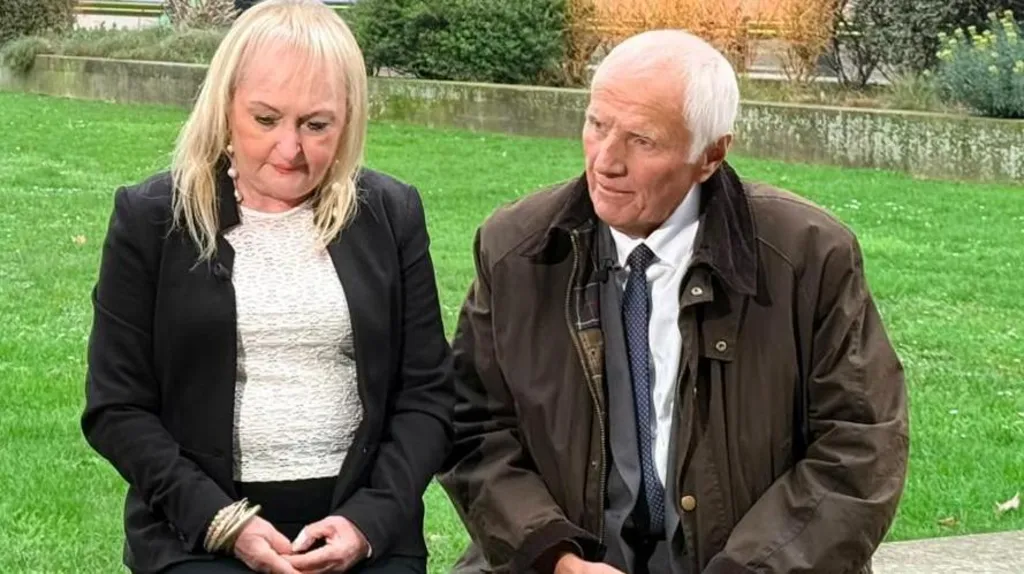 Jenni Hicks, who has blonde hair and is wearing a dark suit jacket over a white top, and Trevor Hicks, who has white hair and is wearing a grey suit and white shirt under a dark greenish brown overcoat, sit on a low wall in front of a patch of green grass and speak to a journalist who is behind the camera