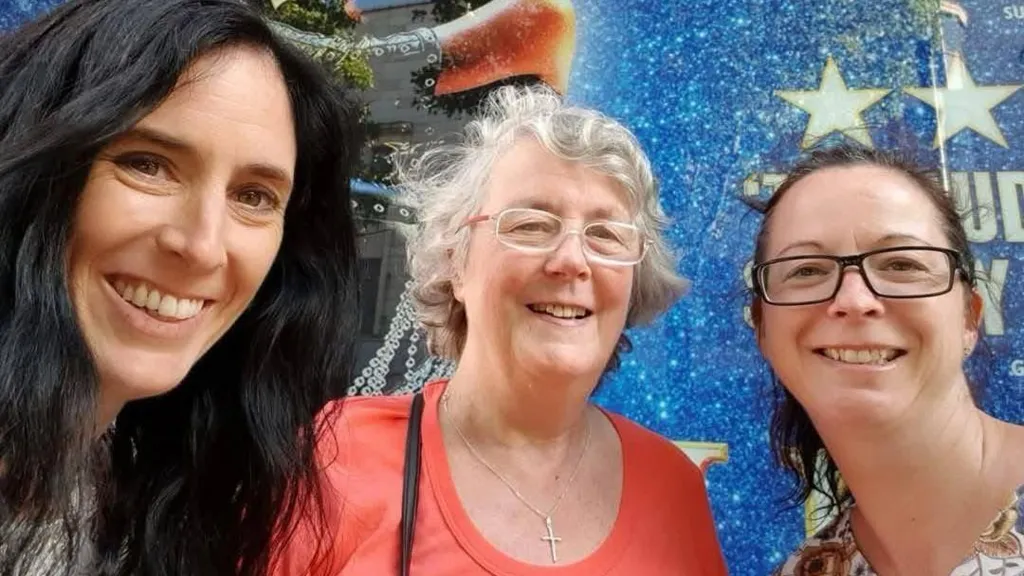 Three women smile in a selfie against a blue glittery backdrop. The woman on the left has long loose black hair, the one in the middle has white short curly hair and glasses and is wearing an orange scoop-neck t-shirt. the woman on the right has black hair tied back and dark-framed glasses.