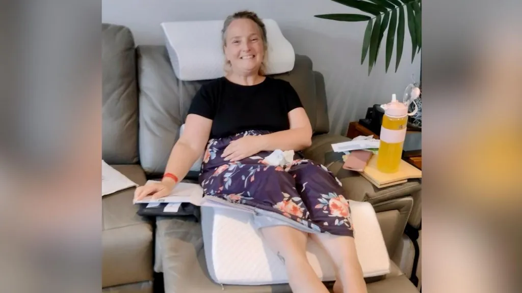 A woman in a black top and floral purple skirt is sitting on a grey sofa with her legs raised and a triangular white foam pillow under her knees.