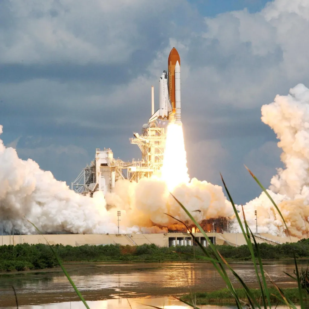 Space Shuttle during launch at Cape Canaveral in Florida. A white shuttle is attached vertically to an orange rocket with large white booster engines. Flames are firing out of the rocket engine and there re large amounts of white smoke as the rocket is just about to lift off. In the foreground there is a a lake.