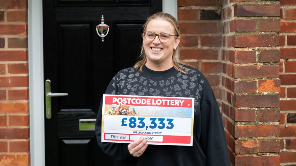 Woman smiling in front of a door holding £83,333 postcode lottery ticket