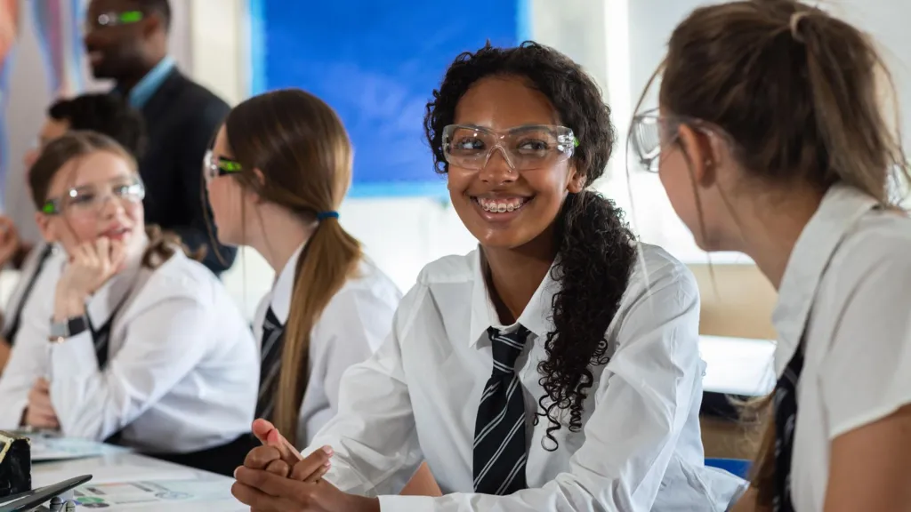 A medium over-the-shoulder shot of a group of teenage students, dressed in matching school uniforms consisting of white shirts and striped ties, are seated around a long lab table in a bright and modern science classroom. The students appear engaged and cheerful, smiling and chatting with each other.