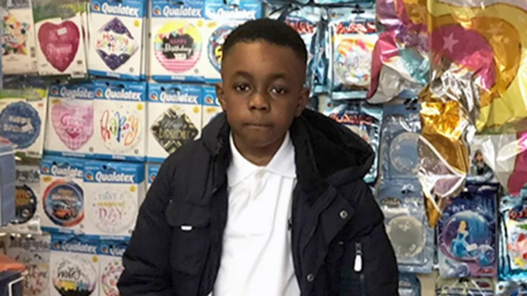 Daejaun, pictured as a young boy, stands in a party‑supply store in front of a wall of colourful packaged balloons. The display features various foil balloon designs, including “Happy Birthday,” hearts, stars, and character-themed shapes. Larger star and moon balloons hang to the right. Daejaun wears a dark jacket over a white shirt and is centred in the frame.
