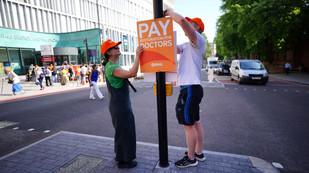 A man and a woman wearing orange BMA caps attach at Pay Doctors placard to a post on a traffic island outside Bristol Royal Infirmary. The entrance of the hospital, with pickets and media outside, is visible in the background on the left side of the shot, while cars queue on the right hand side. 