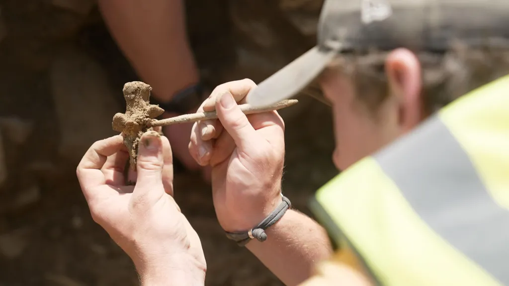 Close-up of an archaeologist’s hands carefully cleaning a small bone fragment. They are holding the bone in one hand and using a thin wooden tool to scrape away the soil that it is covered with. The archaeologist is wearing a yellow high-visibility vest and a cap.