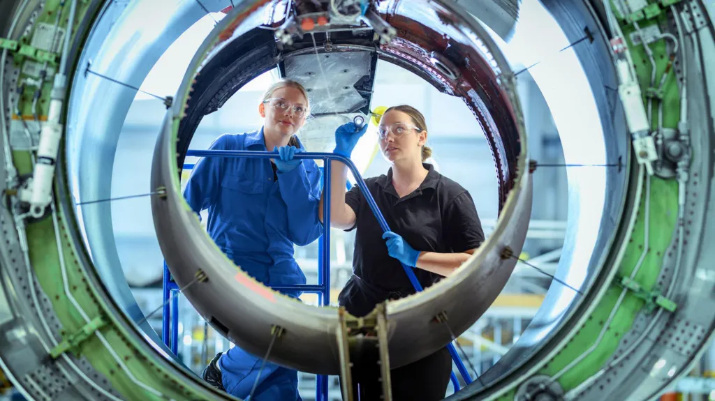 A female apprentice, in blue overalls, is shown jet engine housing by an instructor, wearing black overalls - they are both wearing safety goggles and blue gloves 