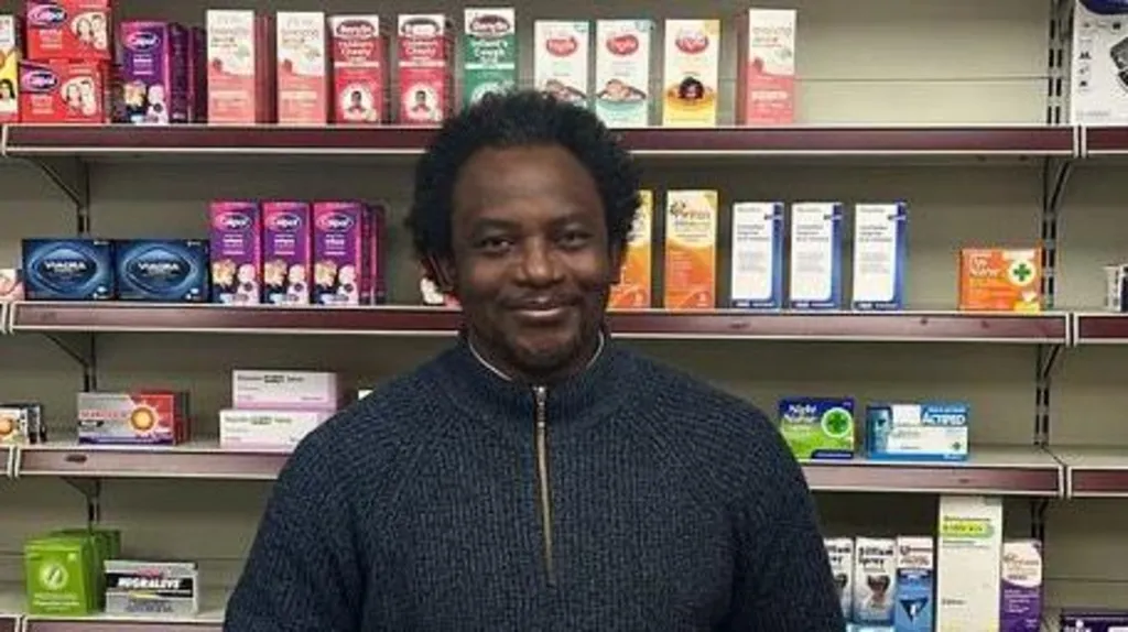 Tunde Olawoye stood behind the counter at his pharmacy wearing a blue fleece jumper. There are shelves of drugs and medicine behind him.