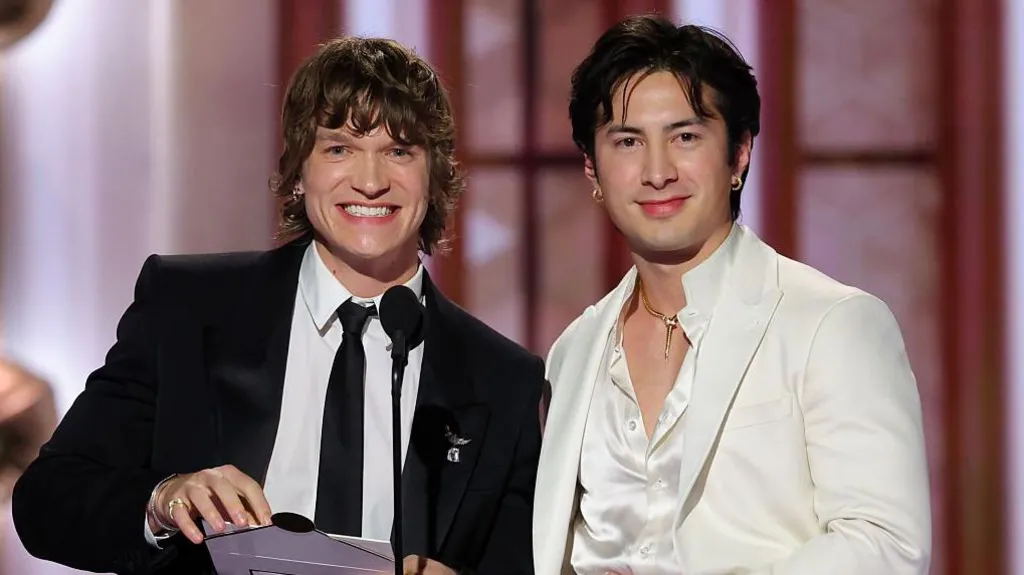 Connor Storrie, smiling and holding an envelope, wearing a dark suit and tie, and Hudson Williams in a white open necked shirt a white suit coat, on stage at the Golden Globes 