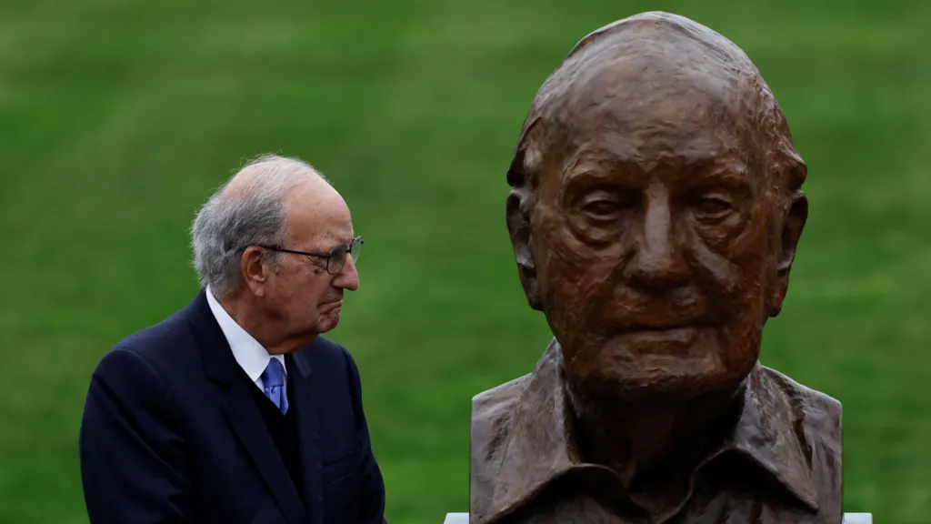 George Mitchell, with short grey hair, wearing glasses, a white shirt, purple tie and navy suit. He is looking to the right at a bust depicting himself.