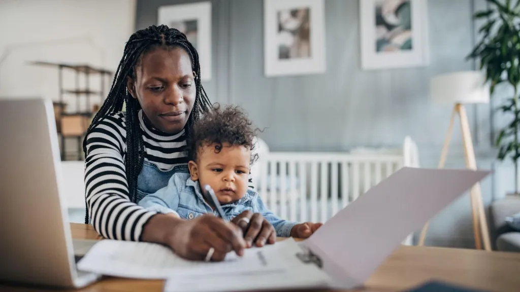 A mother working from home with a laptop, writing notes on a piece of paper with her son on her lap.
