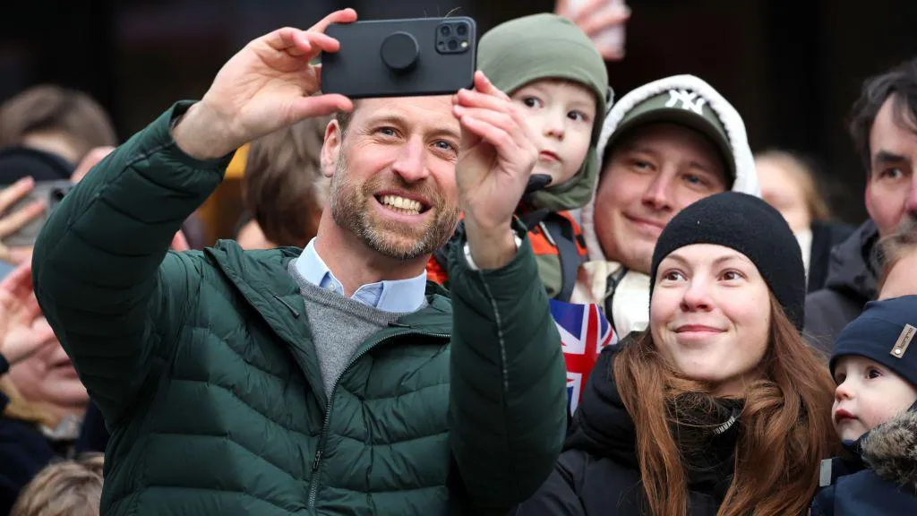 Prince William holds a phone up to take selfies with people in Tallinn on his visit to Estonia in March 2025. He is wearing a green padded jacket, with a grey jumper and blue shirt underneath. Members of the public are crowded around.