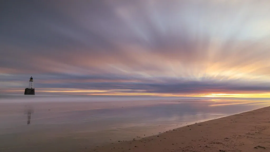 A wide sandy beach at sunset, with soft pink and purple clouds streaking across the sky. A lone lighthouse stands in the distance on the left, reflected in the calm water.