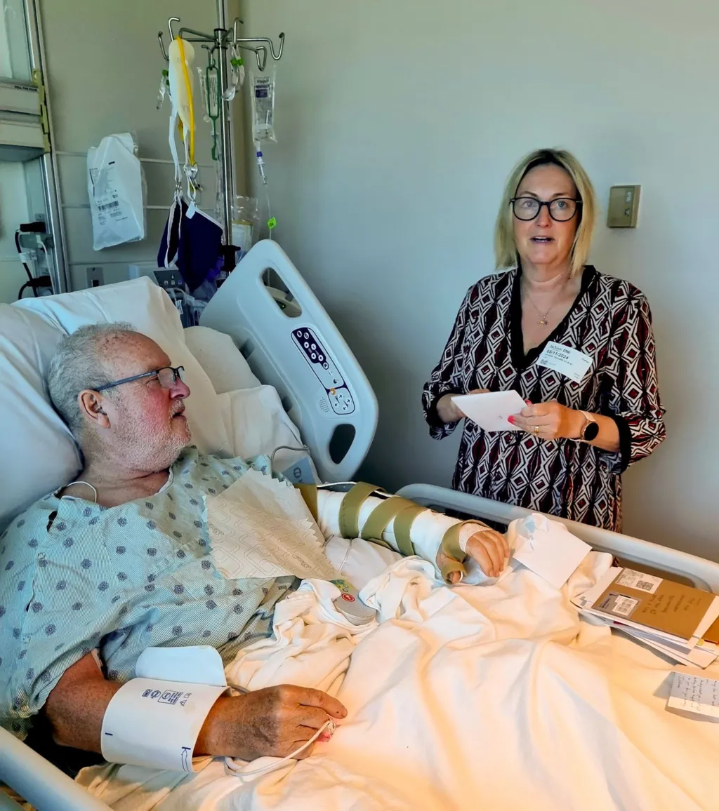 Peter - left arm bandaged up- lying in hospital bed, with wife Joanna by his side.  