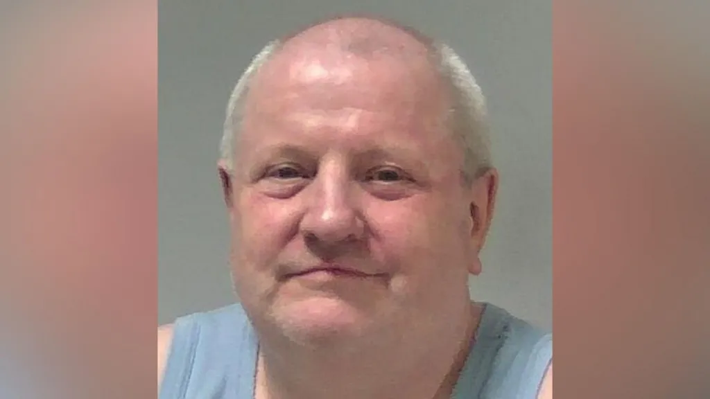 A custody photo of Steve Wright who is smiling at the camera. He is wearing a light blue vest and is bald with some grey hair around the side of his head.