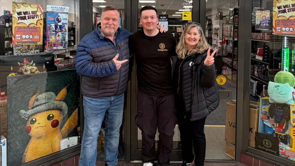Shop owner Alex Badham outside his gaming shop, he's smiling and stood next to a man and a women. Alex has short dark hair, is cleanly shaven and is wearing a black t-shirt and trousers. The woman next to him is making a piece symbol with her hands and smiling, while the man on Alex's other side is pointing at Alex and smiling. 