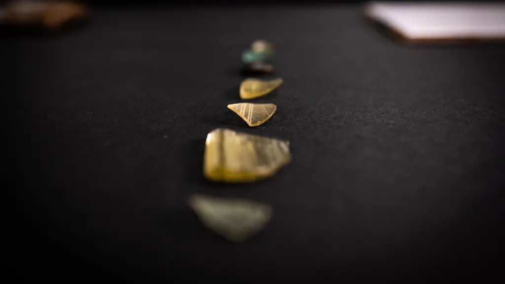 Several fragments of glass arranged in a straight line on a dark surface. They vary in size and shape, and some having a yellow tint and others a green and blue hue. One shard is finely etched with a pattern.