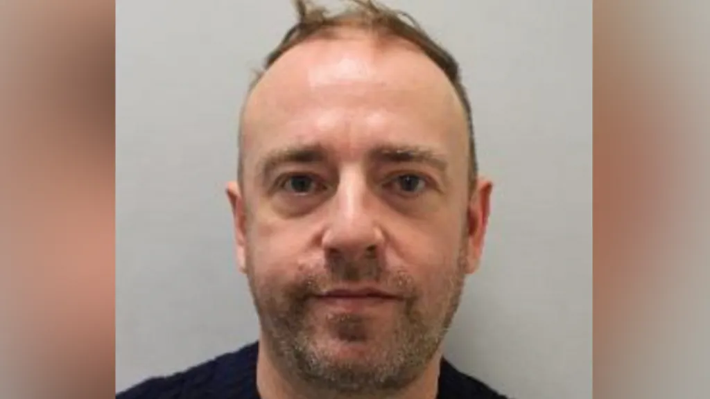 A mug shot of Gunnery who has brown hair and facial stubble and is wearing a black jumper